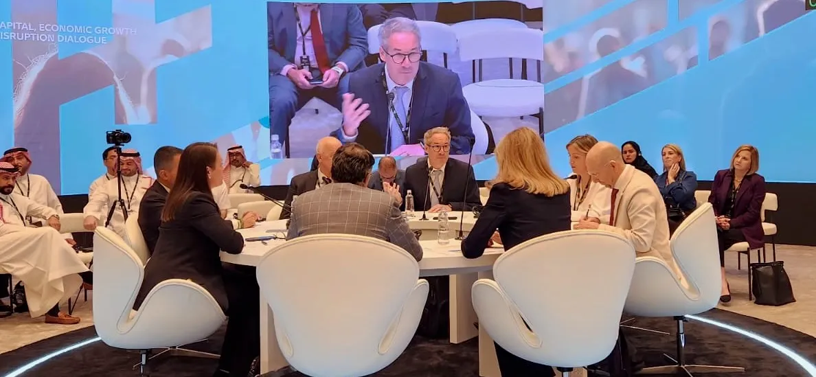 Andrew Kilshaw speaking at a round-table session, shown on the room’s large screen above delegates seated around a white table.