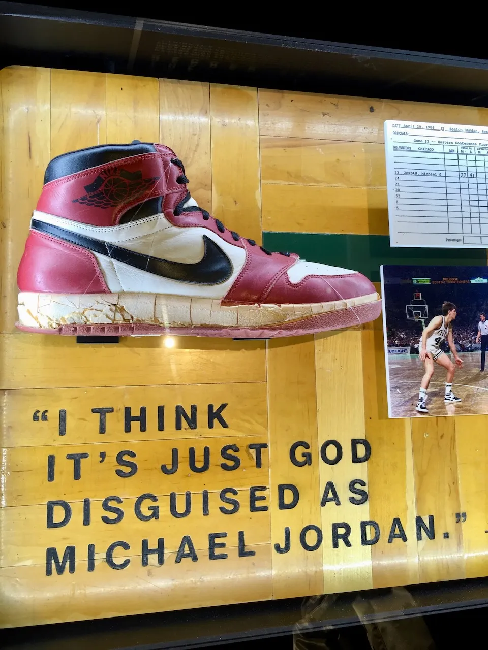 An Air Jordan 1 in a glass display case, its sole cracked with age, beside a box score from a game at Boston Garden on 20 April 1986 and a photograph of Larry Bird. A quotation on the case reads: I think it’s just God disguised as Michael Jordan.
